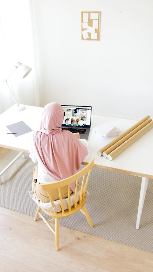 A Woman in Pink Hijab Sitting on a Wooden Chair while Using Her Laptop