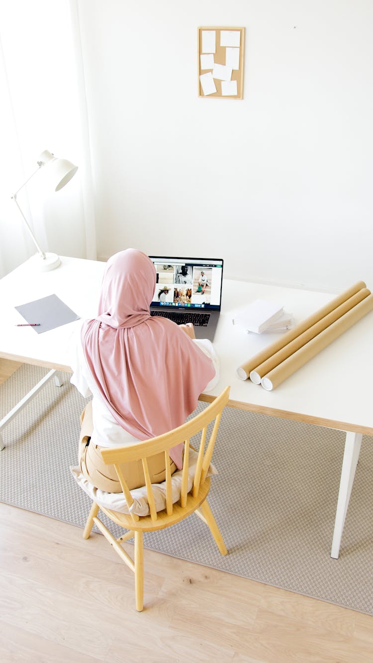 A Woman In Pink Hijab Sitting On A Wooden Chair While Using Her Laptop