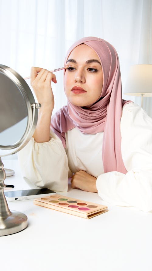 A Woman in Pink Hijab Applying Eyeshadow while Looking at the Mirror