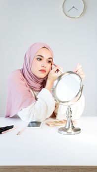 A middle eastern woman in hijab applies makeup at a mirror, embodying beauty and tradition.