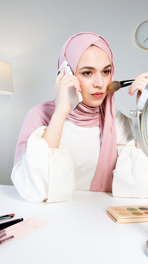 A Woman in Pink Hijab Applying Makeup while Talking on the Phone