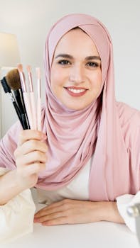 A cheerful woman in a pink hijab holds makeup brushes while smiling indoors.