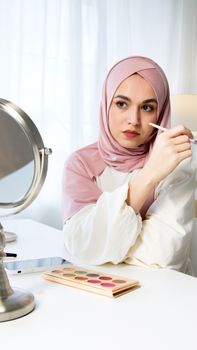 Woman in hijab applying makeup at home using a palette and mirror.
