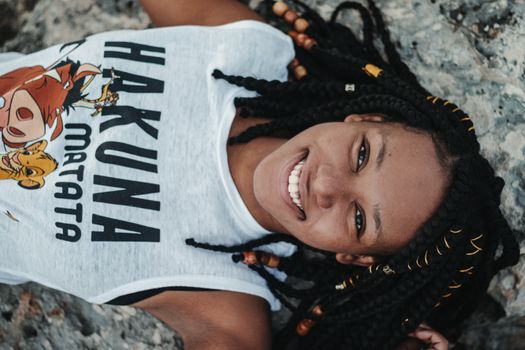 Top view of young cheerful black female in stylish clothes with inscription and image looking at camera while lying on stone