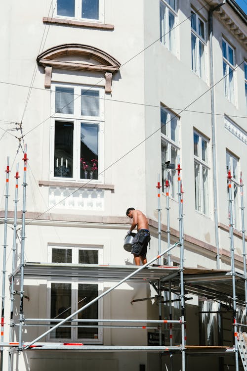 A Shirtless Man Standing on a Scaffolding Near the Building with Glass Windows