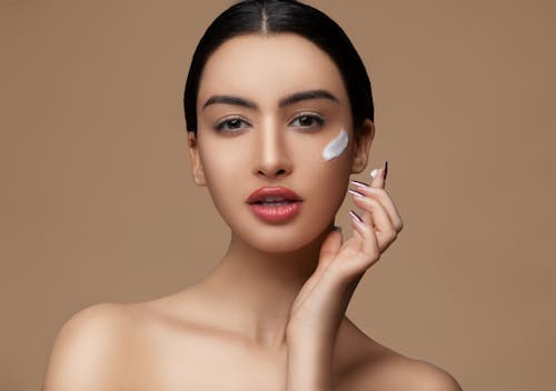 Crop sensitive female with makeup and red lips applying cream on delicate face skin while looking at camera on beige background