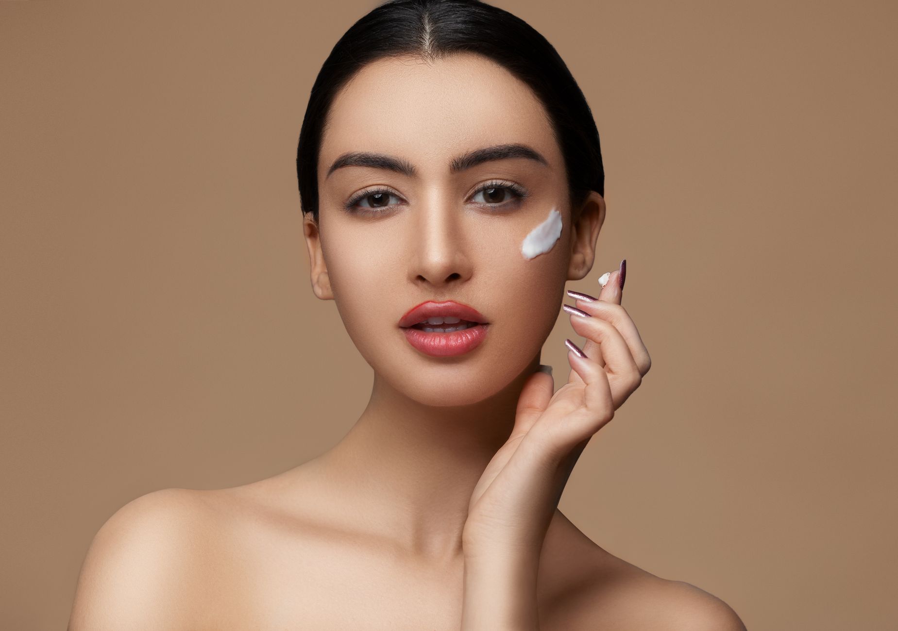 Crop sensitive female with makeup and red lips applying cream on delicate face skin while looking at camera on beige background