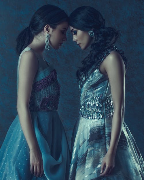 Free Side view of young women with closed eyes and makeup touching foreheads while standing in stylish dresses and earrings Stock Photo
