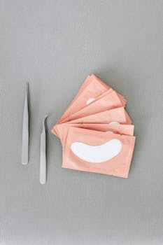 Metal tweezers and eye patches for eyelash extensions on a gray surface, offering a minimal aesthetic.