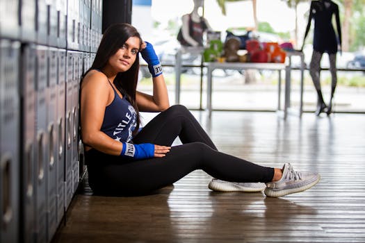 Woman in activewear sitting by gym lockers with a thoughtful expression, wearing blue boxing wraps.