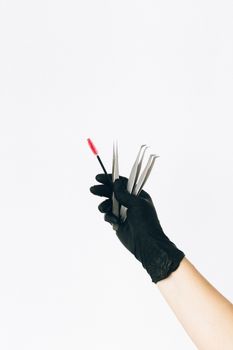 A black gloved hand holds multiple tweezers and a brush against a white background.