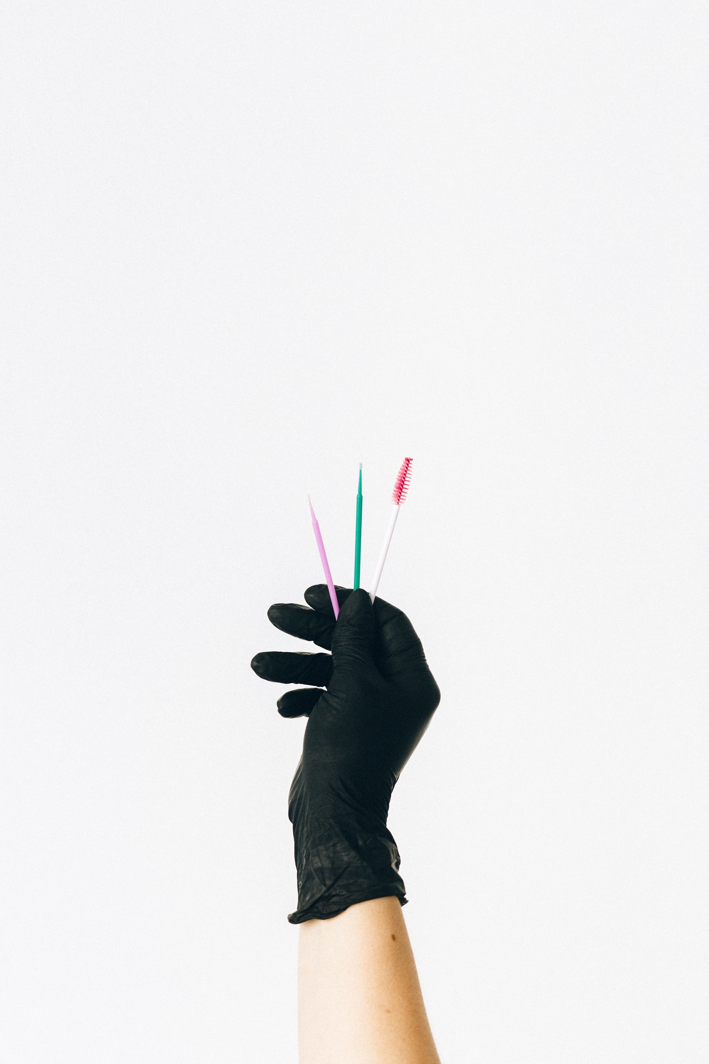 A black-gloved hand holds three slim pink, green and white cosmetic applicator tools against a plain white background.