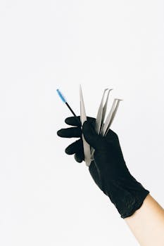 A hand in black glove holding tweezers and mascara brush against a white backdrop.