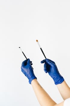 Two hands in blue latex gloves holding makeup brushes on a plain background.