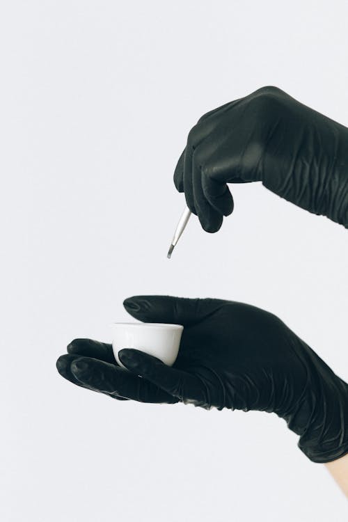 Hands of a Person in Black Gloves Holding Makeup Brush and White Bowl