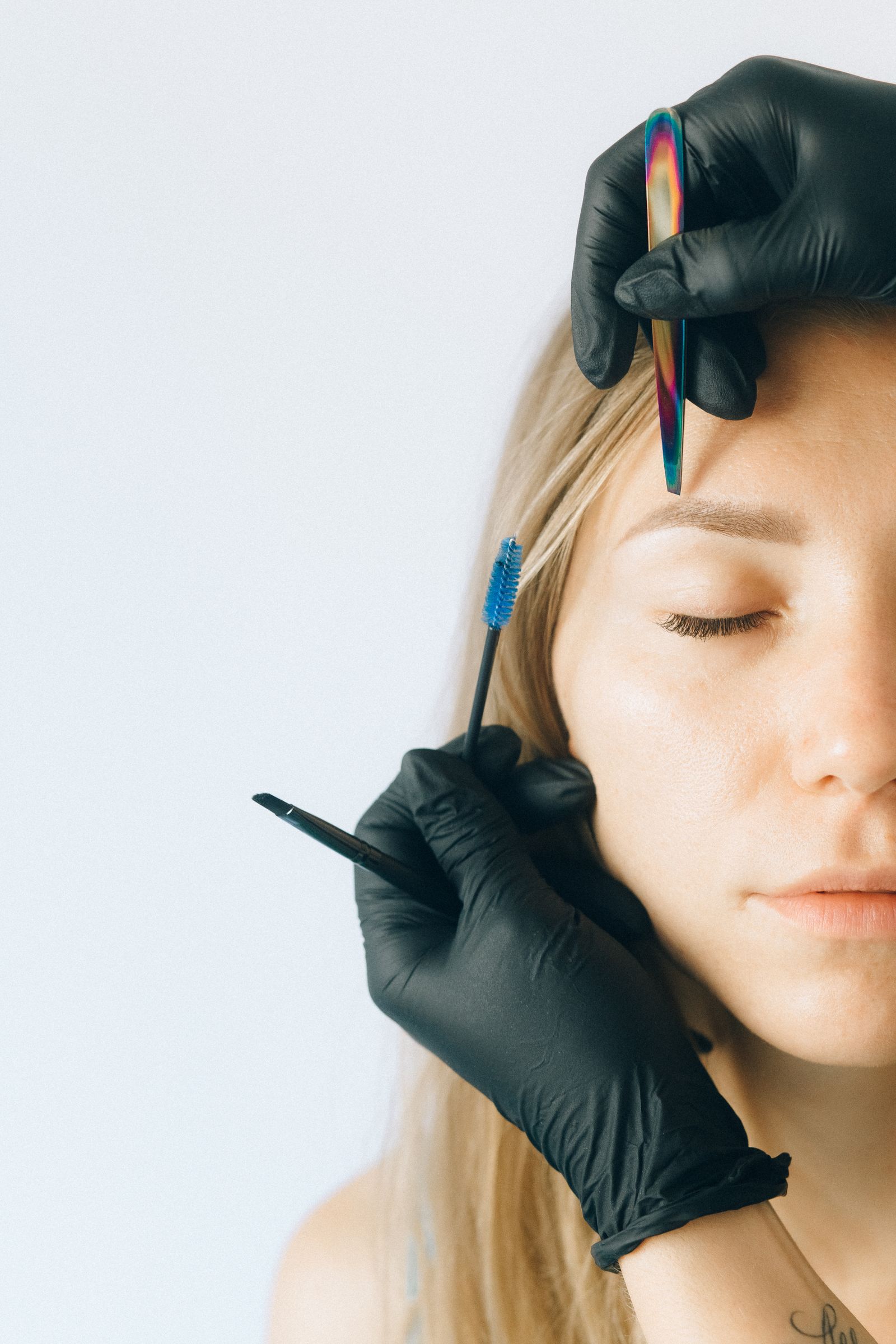 Close-up of a gloved technician holding a multicoloured precision tool above a client's eyebrow, with a blue spoolie and angled brush nearby.