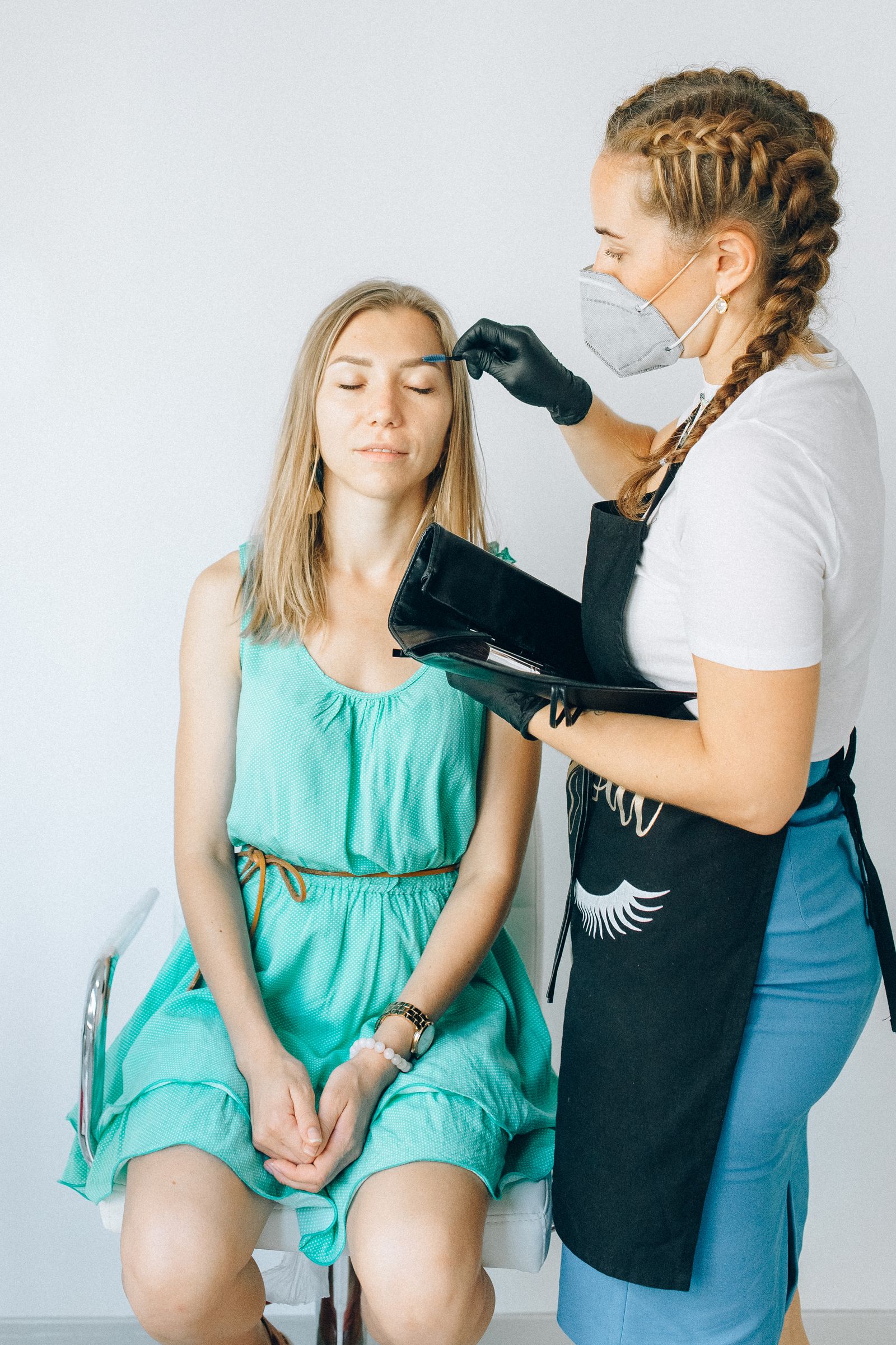 A masked practitioner wearing gloves uses a small tool near a seated client's eyebrow while holding a treatment case.