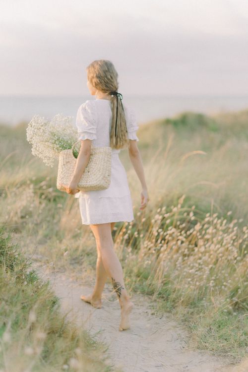 Free A woman in a white dress walks barefoot along a scenic coastal path holding flowers. Stock Photo
