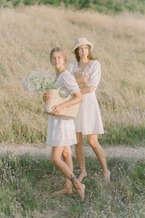 Two Women in White Dress Holding Basket on Green Grass Field