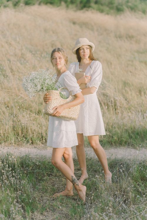 Free Two women in white dresses carrying flowers walk barefoot in a sunlit field. Stock Photo