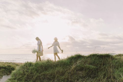 Free Two women walking on grassy dunes at the beach during a sunny summer day, capturing a bohemian vibe. Stock Photo