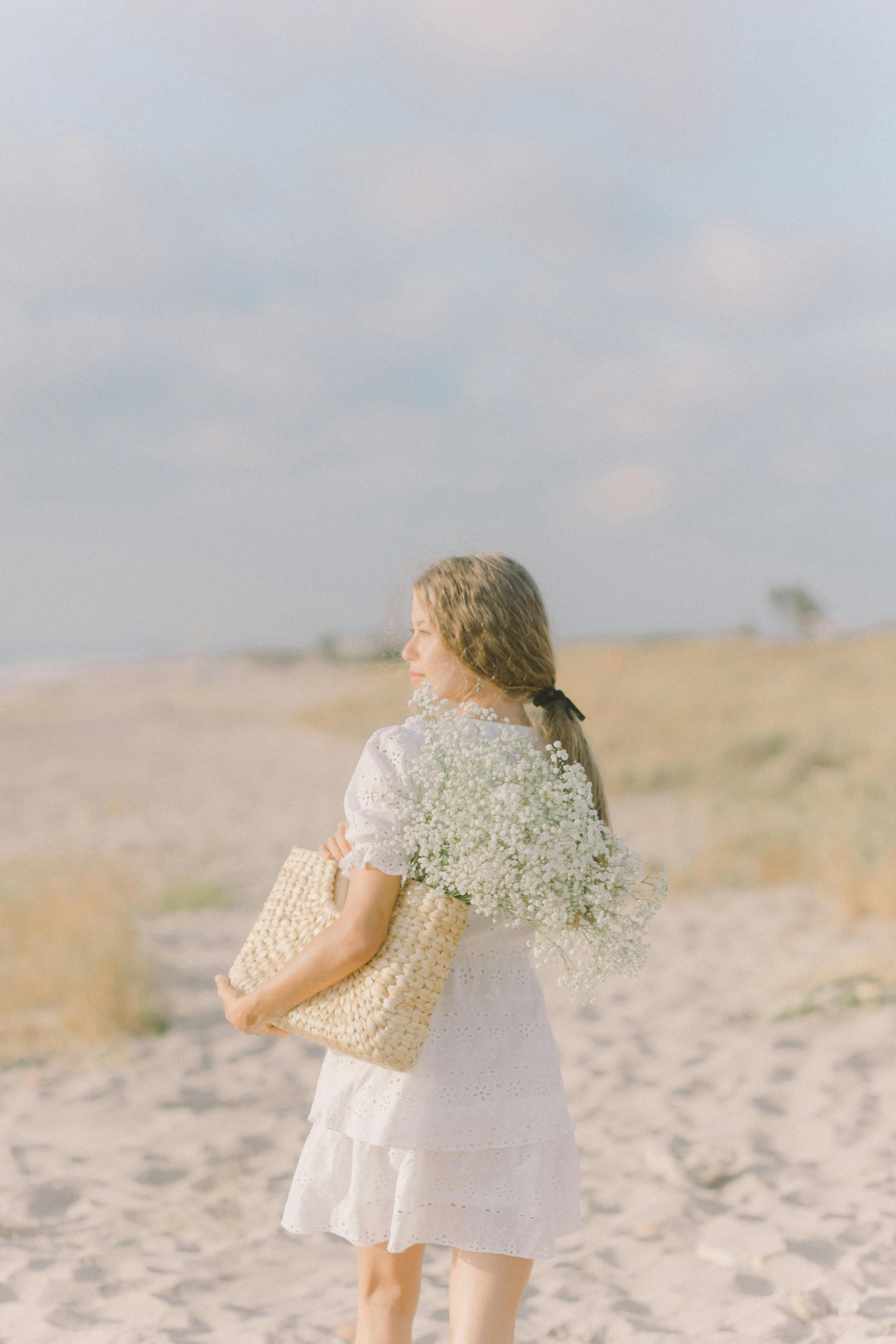 一位身穿白色连衣裙的年轻女子在阳光明媚的海滩上手握一篮子鲜花，捕捉到宁静的夏日时刻