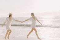 两名身穿白色连衣裙的女子在海滩上牵着手微笑，享受着阳光明媚的一天