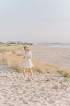 A young woman in a white dress and hat enjoys a sunny day on a sandy beach.