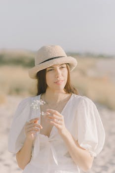 阳光明媚的海滩上，一名头戴草帽、身穿白色连衣裙的年轻女子手拿小白花站在那里