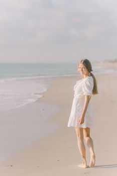 一名年轻女子身穿白色连衣裙，赤脚走在阳光明媚的海滩上，享受着宁静的夏日