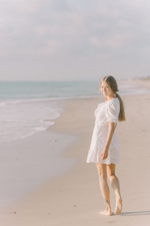 Free A young woman in a white dress walks barefoot on a sunny beach, enjoying a peaceful summer day. Stock Photo