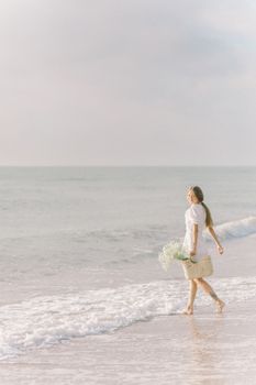 Serene scene of a woman in a white dress walking along the beach with a basket on a sunny day.
