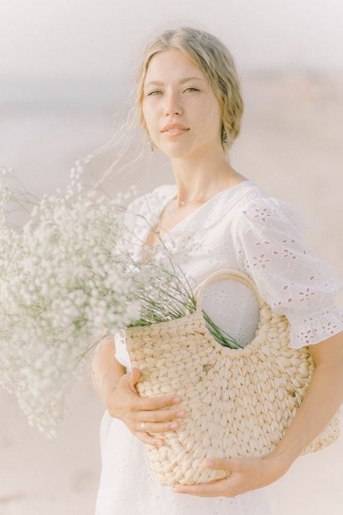 Free A young woman in a white dress holds a basket filled with delicate flowers on a serene beach. Stock Photo