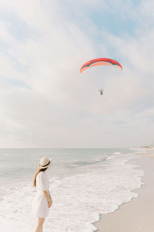 Free Elegant woman in white dress watches a paraglider soar over the tranquil beach. Stock Photo