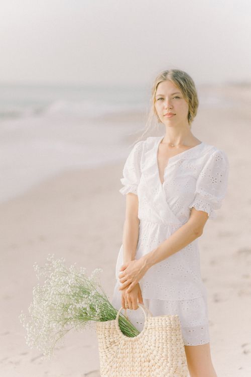 Free Young woman in a white dress holding basket with flowers on a serene beach. Stock Photo