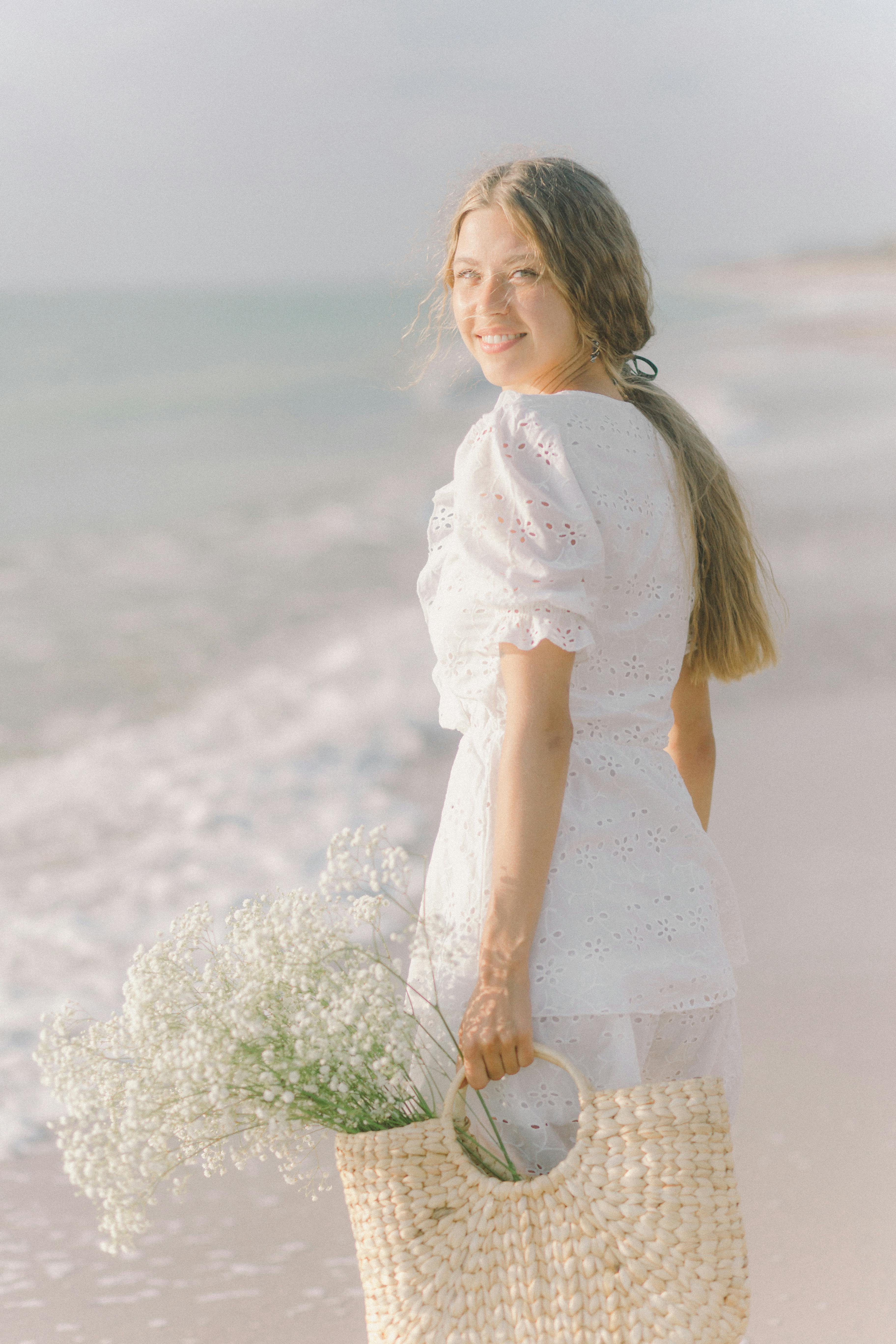 夏天，一名身穿白色连衣裙的年轻女子在海滩边微笑，篮子里拿着鲜花