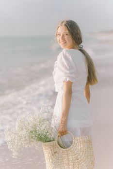 夏天，一名身穿白色连衣裙的年轻女子提着装满鲜花的篮子，在海滩边微笑