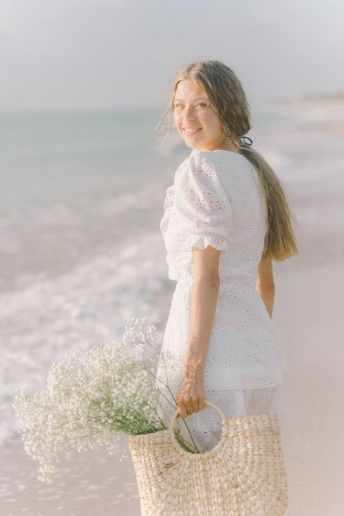 Free A young woman in a white dress smiles by the beach during summer, holding flowers in a basket. Stock Photo