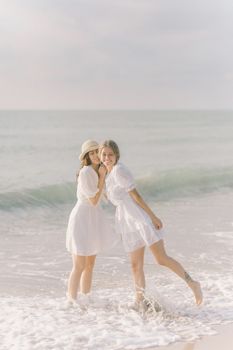 两名身穿白色连衣裙的女子在海滩上享受阳光明媚的一天，欢乐与友谊洋溢其间