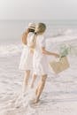 两名身穿白色连衣裙的女子，提着一个装满鲜花的篮子，沿着阳光明媚的海滩散步，享受着夏天
