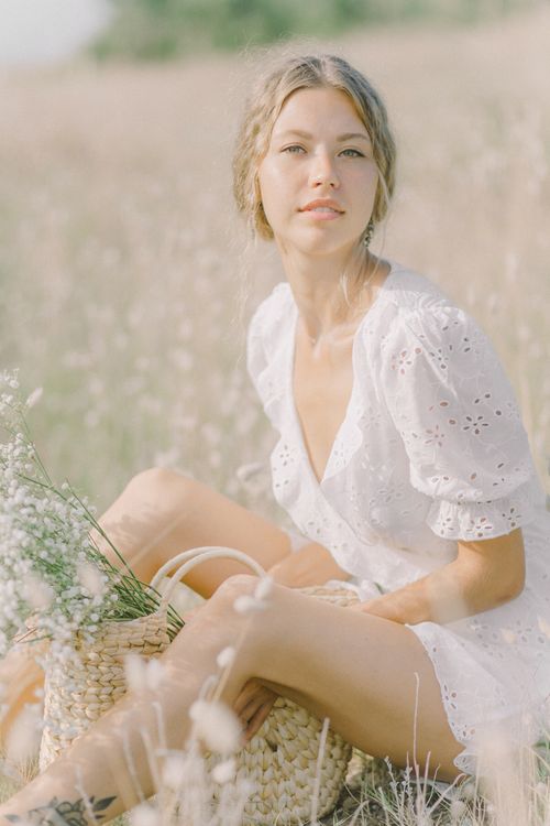 Free A woman in a white dress sitting in a field, holding a basket with flowers. Stock Photo