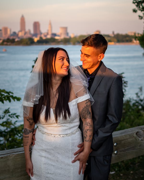 Groom and bride with tattoos standing near water and looking at each other while enjoying wedding celebration in city street