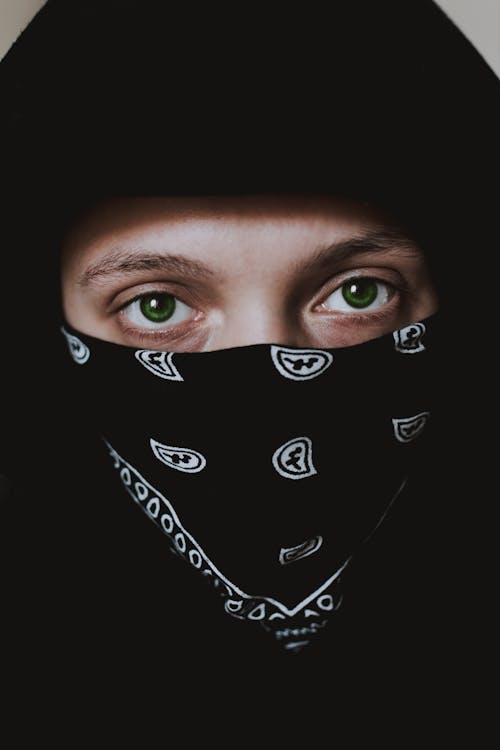Anonymous person with green eyes in black hood and black bandana with white pattern looking at camera