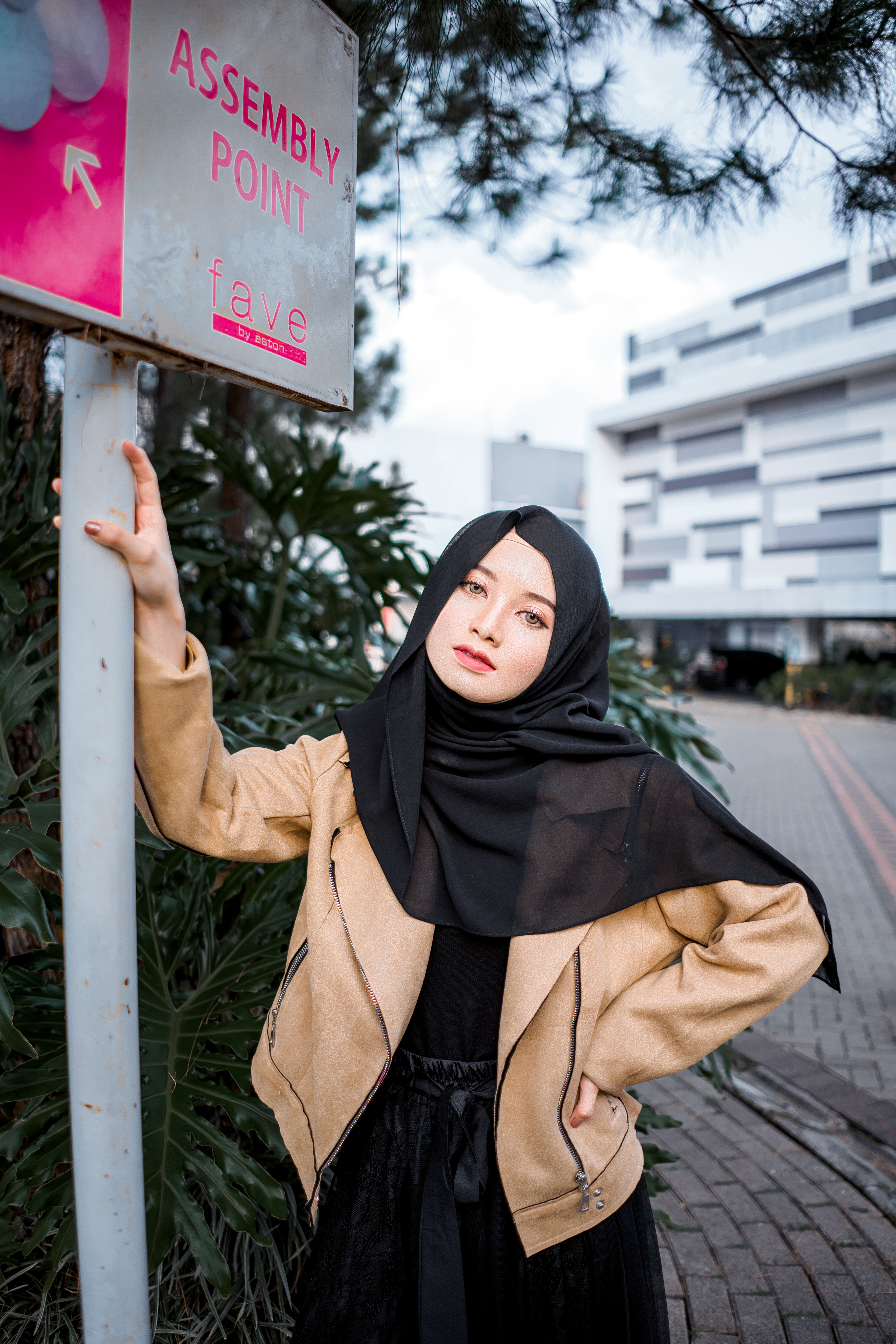 Young ethnic female in trendy outfit and traditional hijab standing on paved street with hand on waist near direction signboard and looking at camera