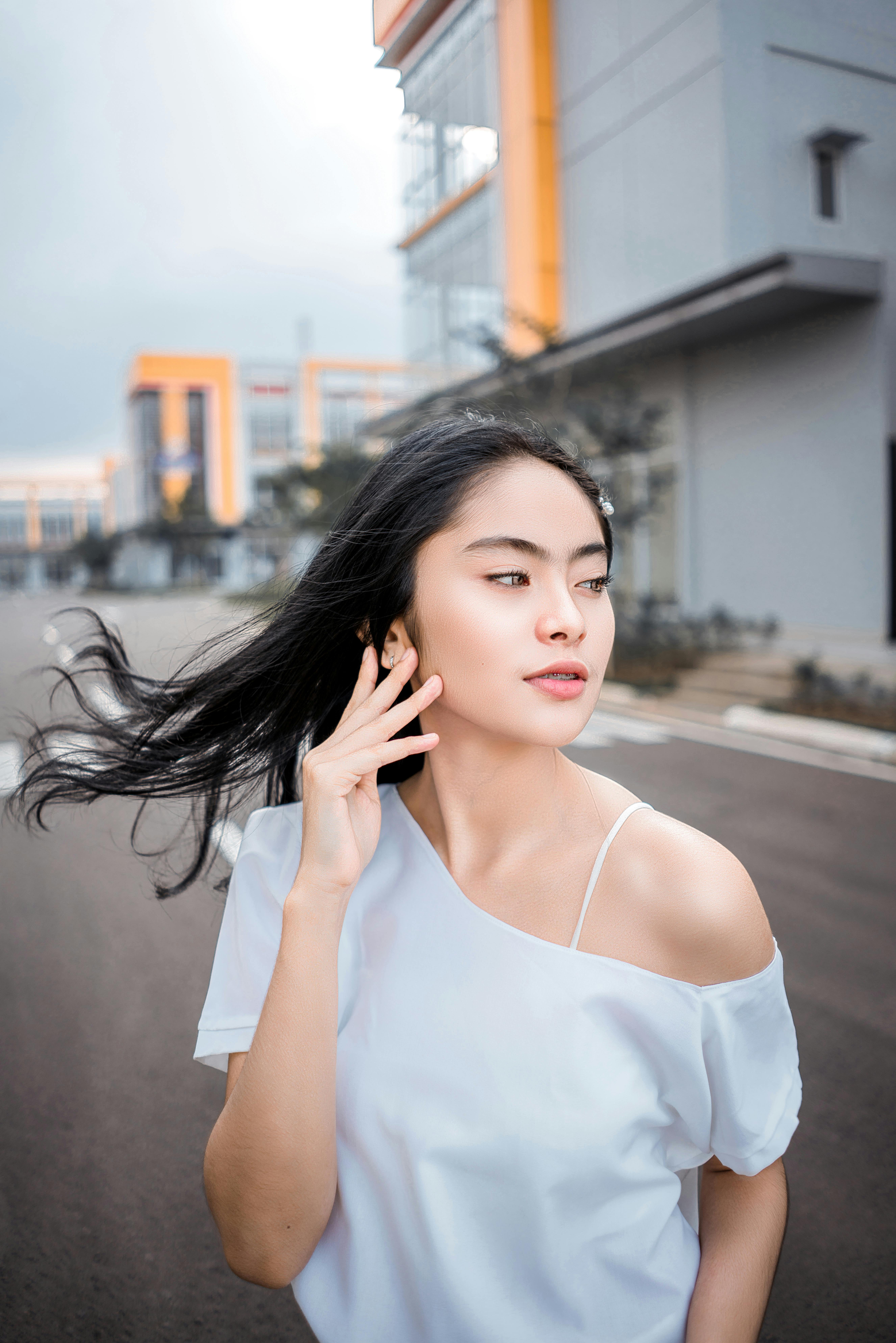 Content young Asian lady with long dark hair in casual clothes touching face and looking away while standing on street near modern buildings