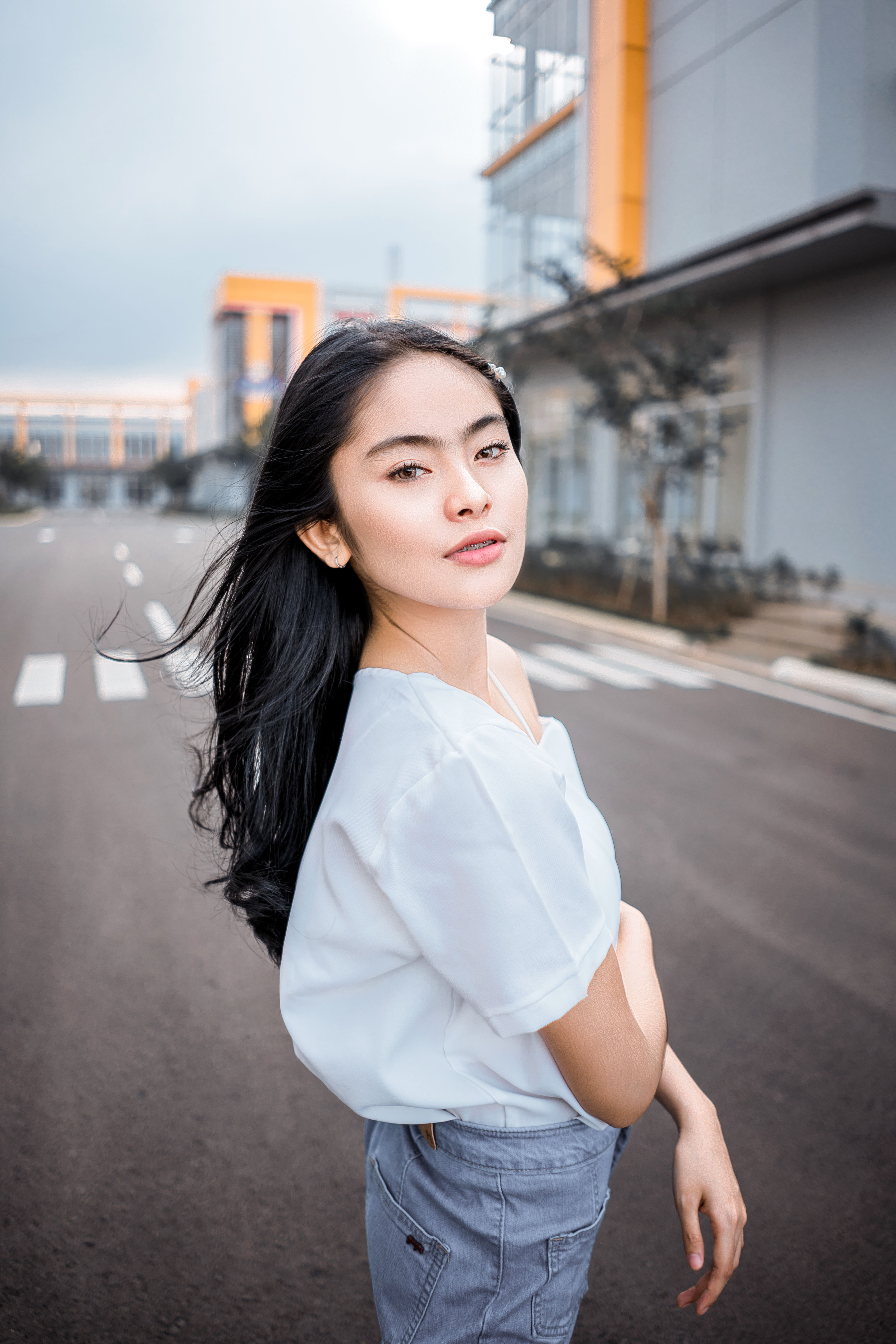 Side view of young stylish Asian female millennial with long dark hair in casual outfit standing on asphalt roadway in city modern district and looking at camera