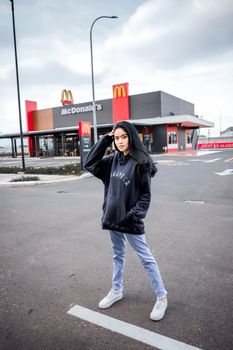Full length of young stylish ethnic female in trendy hoodie and sneakers standing on asphalt parking area near fast food cafe on cloudy day