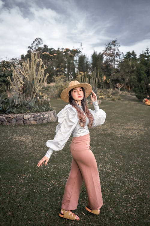 Side view of stylish young female traveler in trendy outfit and straw hat walking along grassy lawn while enjoying holidays in park on cloudy day