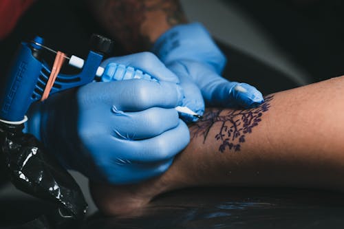 From above of faceless tattooist in blue gloves applying tree tattoo with ink and professional machine on leg of anonymous client in salon
