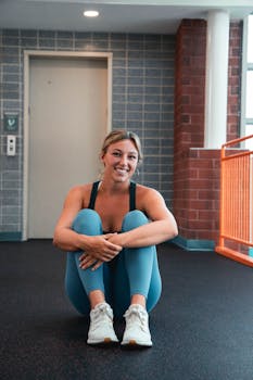 Smiling woman in athletic attire sitting on the floor in a modern sports facility.